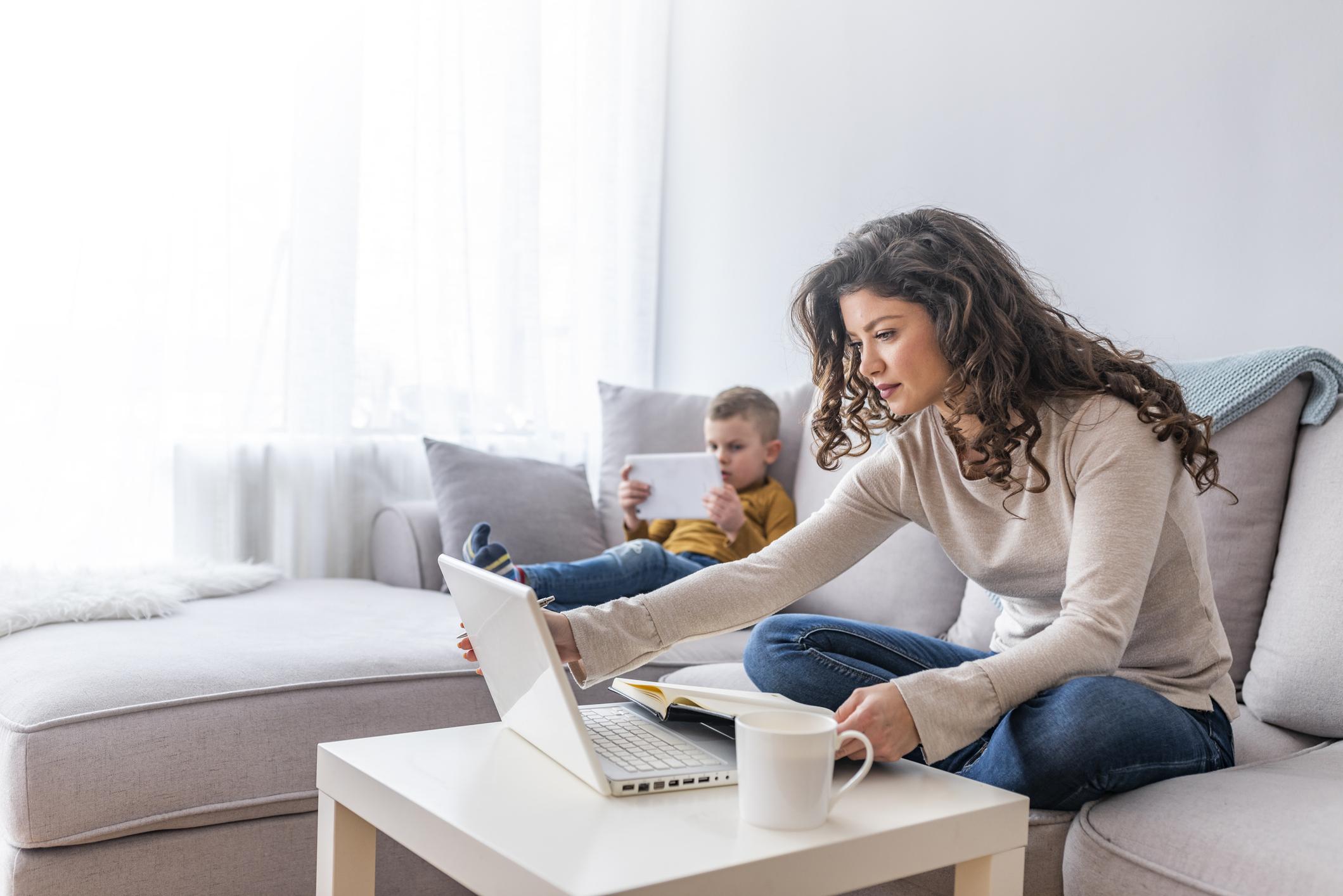 woman sitting with laptop with child in background on tablet